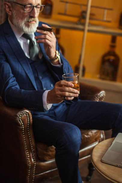 Elderly businessman with glass of whiskey and cigar on dark background - Фото, зображення