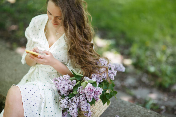 The girl in white dress holds in her hands a wicker basket with flowers. Basket with lilacs. - Foto, imagen