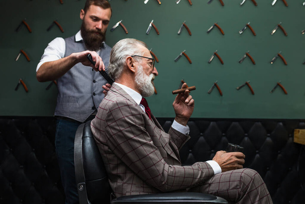 Bearded old businessman sitting at barber shop in chair choosing haircut design - Foto, immagini