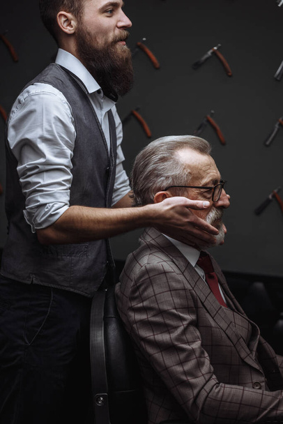 Bearded old businessman sitting at barber shop in chair choosing haircut design - Foto, afbeelding