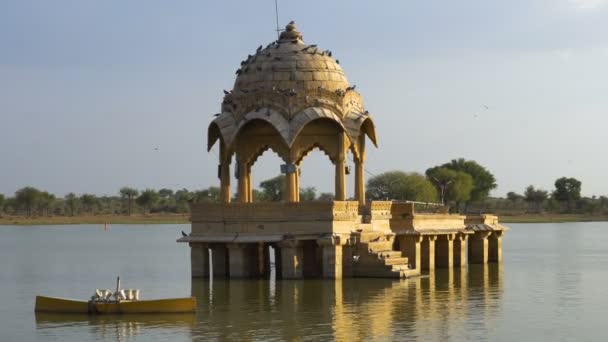 Estructuras hechas de piedra amarilla en medio del cuerpo de agua, lago Gadisar en la hermosa ciudad de Jaisalmer, construido artificialmente para la conservación del agua. Le dispararon a Jaisalmer, Rajastán, India. Fondo cielo azul. - Metraje, vídeo