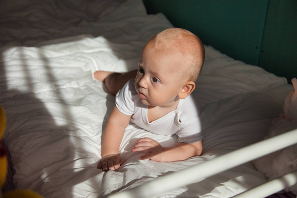 El bebé de 6 meses se acuesta boca abajo y muestra emociones. Adorable chico rubio se encuentra en la cama - Foto, Imagen