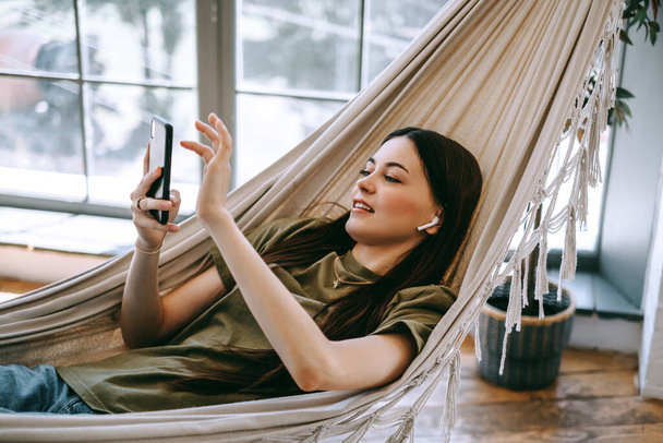 Atractiva mujer joven feliz relajarse en el hogar, sentado en la hamaca en la sala de estar, utilizando el teléfono inteligente, hablando en el chat de vídeo, saludando. Foto de alta calidad - Foto, Imagen