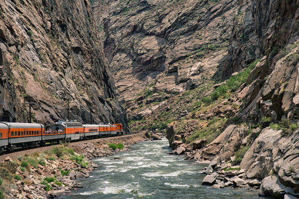 Juna ratsastaa syvällä kuninkaallisessa rotkossa Arkansas-joen varrella Coloradossa. Appelsiinimoottori ryntää sinisen veden viereen kallioiden ympäröimänä. - Valokuva, kuva