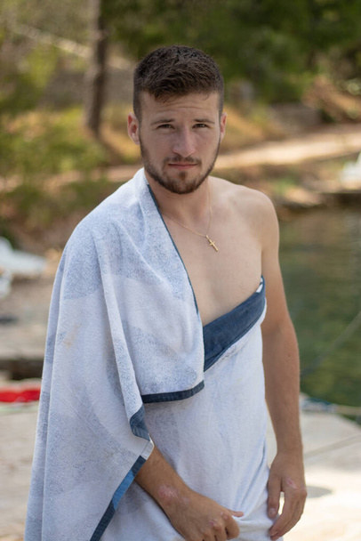 Adult man wearing a towel as tunic around his waist looking at the camera with the adriatic sea in the background - Photo, Image