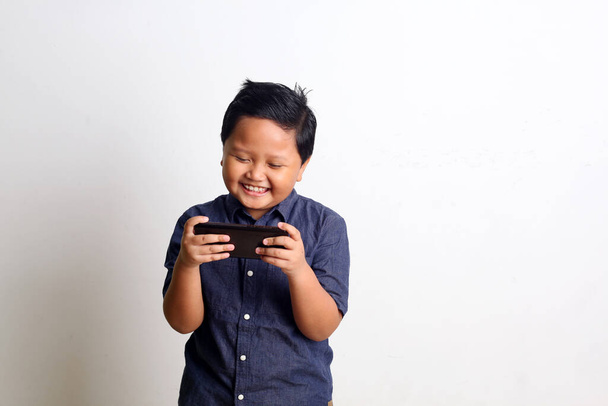 Niño asiático de pie y jugando en el teléfono inteligente. Aislado en pared blanca - Foto, imagen