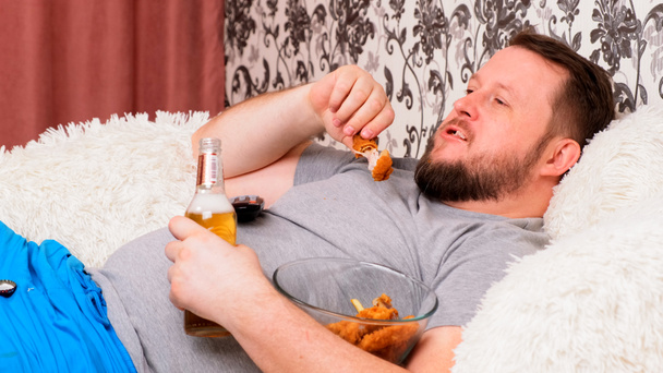 Dicker Mann trinkt Bier und isst ungesundes Essen Chicken Wings, gelangweilt vor dem Fernseher auf der Couch. Das Konzept der Unterernährung, Quarantäne zu Hause, Alkoholismus. - Foto, Bild