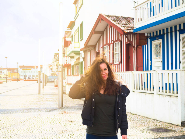  una giovane donna in giacca con i capelli lunghi cammina per le strade con case a strisce colorate, Costa Nova, Aveiro, Portogallo - Foto, immagini