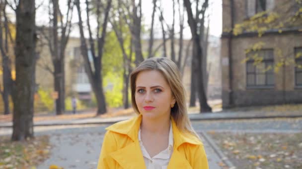 黄色のマントを着た女性、秋の公園に立つ白いブラウス、カメラを見て - 映像、動画