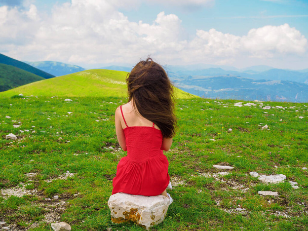 A young woman in a red dress sits on a rock with her back to the camera in the middle of a green valley in Castelluccio di Norcia, Umbria, Italy. - Fotó, kép