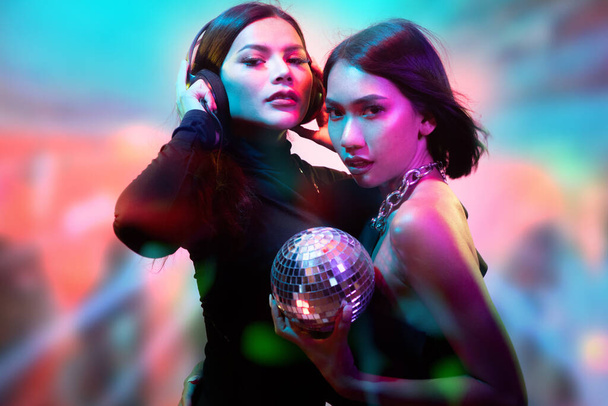 Two young flirting cute Asian women in black bodysuits with headphones and small disco ball posing on colorful bright lights  - Photo, Image