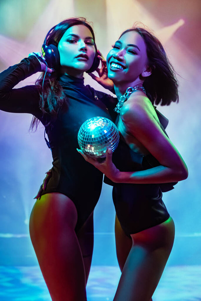 Zwei junge flirtende süße asiatische Frauen in schwarzen Bodys mit Kopfhörern und kleiner Discokugel posieren auf bunten Lichtern  - Foto, Bild