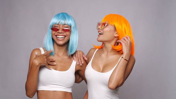 Portrait of two beautiful happy Asian women friends in bright blue and orange wigs and sunglasses posing over gray background - Photo, Image