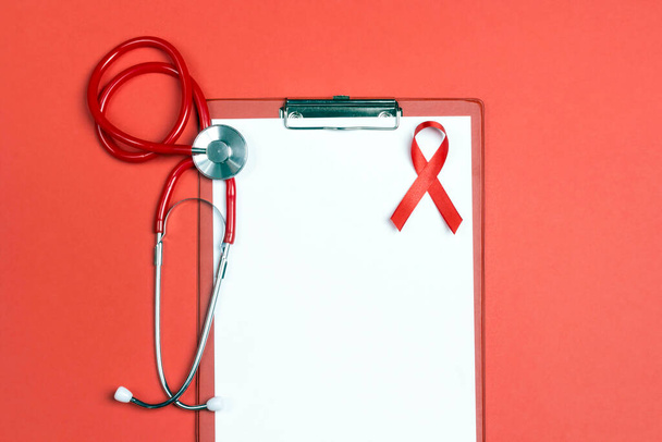 Κενό ιατρικό φάκελο με κόκκινη κορδέλα ως σύμβολο της ευαισθητοποίησης του AIDS και στηθοσκόπιο σε κόκκινο φόντο. 1η Δεκεμβρίου Παγκόσμια Ημέρα κατά του AIDS. - Φωτογραφία, εικόνα