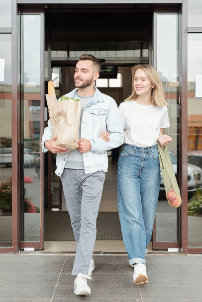Pozytywna młoda para w luźnych strojach, niosąca papierowe torby, wychodząc razem z supermarketu - Zdjęcie, obraz
