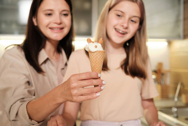 Happy young woman showing yummy homemade icecream in waffle cone decorated by her teenage daughter with almond nuts - Photo, image