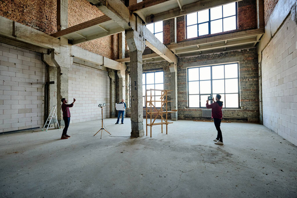 Δύο νέοι εργολάβοι φωτογραφίζουν ημιτελή δομή μέσα στο κτίριο, ενώ ο συνάδελφός τους κοιτάζει το σκίτσο του σχεδίου - Φωτογραφία, εικόνα