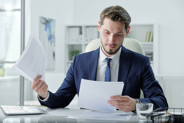 Ocupado joven gerente en traje formal sentado en el escritorio y el examen de documentos de negocios en la oficina - Foto, Imagen