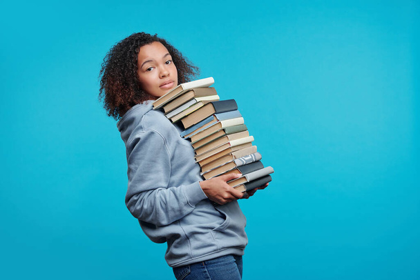 Kıvırcık saçlı siyah kız öğrencinin portresi mavi arka planda bir yığın kitap taşıyor, sınav konseptine hazırlanıyor. - Fotoğraf, Görsel
