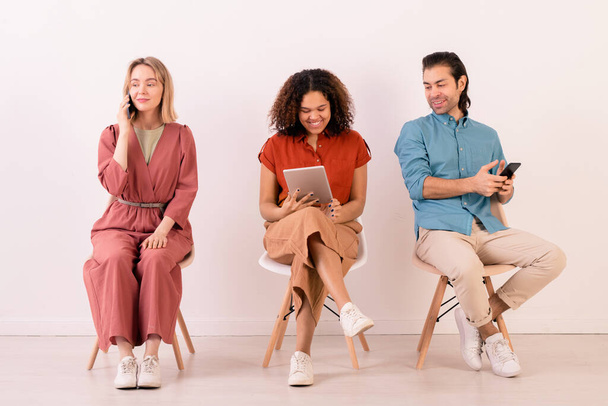 Fila di giovani amici interculturali in casualwear seduti lungo il muro bianco e che comunicano attraverso smartphone e touchpad - Foto, immagini