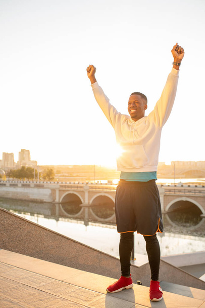 Onnellinen nuori urheilija aktiivisissa vaatteissa pitää kätensä ylhäällä juhliessaan voittoa joenrantaa ja siltaa vastaan aurinkoisena päivänä. - Valokuva, kuva