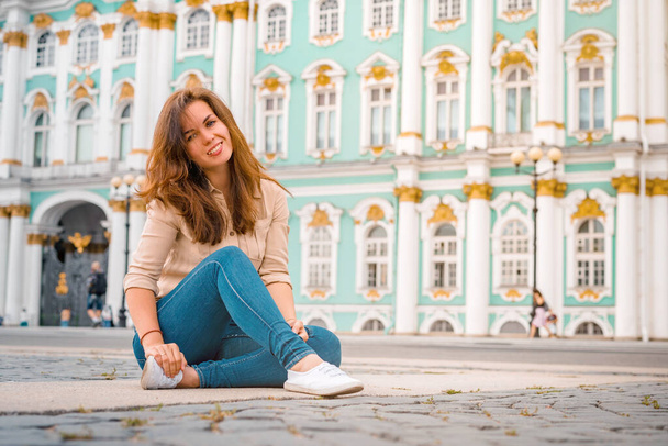 A young woman enjoys near the Winter Palace, Saint Petersburg. Women's tourist walk in the center of Saint Petersburg. A beautiful female traveler is sitting on the asphalt in the St. Petersburg  - Фото, зображення