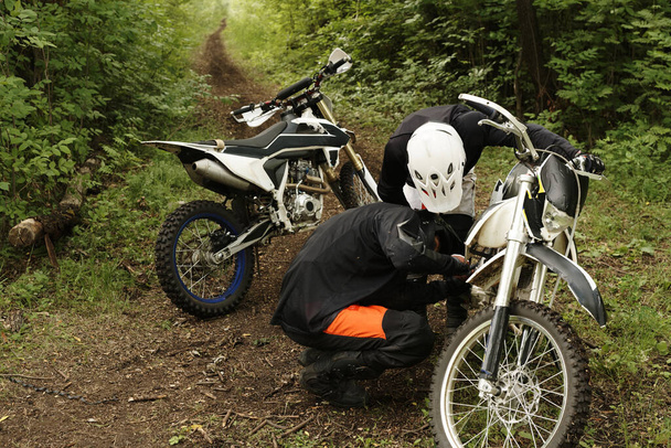 Motociclistas enfocados en cascos revisando motor de motocicleta juntos en el bosque - Foto, Imagen