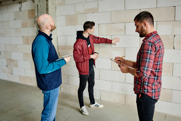 Young contractor with drink touching white brick while standing by wall of unfinished building in front of his colleagues listening to him - Foto, afbeelding