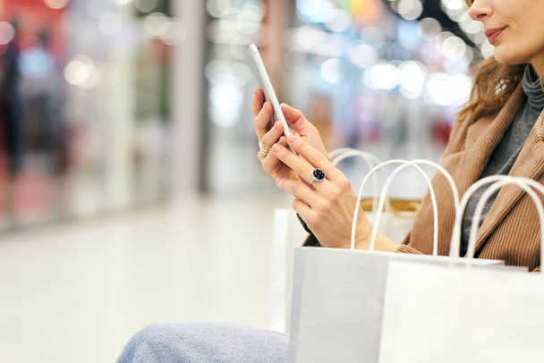 Χέρια των νέων κομψό θηλυκό αγοραστής σε casualwear κοιτάζοντας μέσα από διαφημίσεις στο smartphone, ενώ έχουν ξεκουραστεί μετά τα ψώνια στο εμπορικό κέντρο - Φωτογραφία, εικόνα