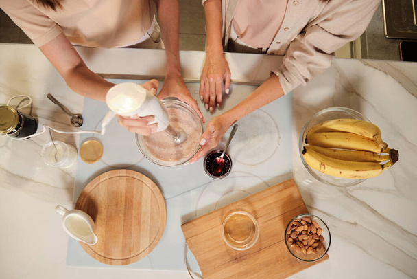 Descripción general de dos mujeres contemporáneas de pie junto a la mesa de la cocina y la mezcla de ingredientes de helado casero con mezclador eléctrico - Foto, Imagen