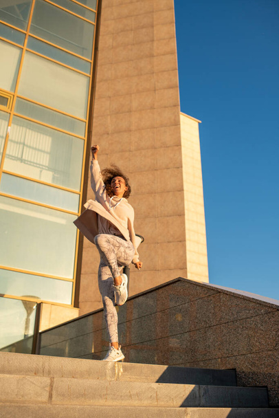 Joven deportista alegre en ropa deportiva manteniendo una pierna doblada en la rodilla mientras está de pie en la parte superior de la escalera contra la arquitectura moderna - Foto, Imagen