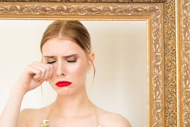 jong boos blonde vrouw met glamoureuze make-up nemen traan uit haar rechteroog achter grote decoratieve frame in de voorkant van de camera - Foto, afbeelding