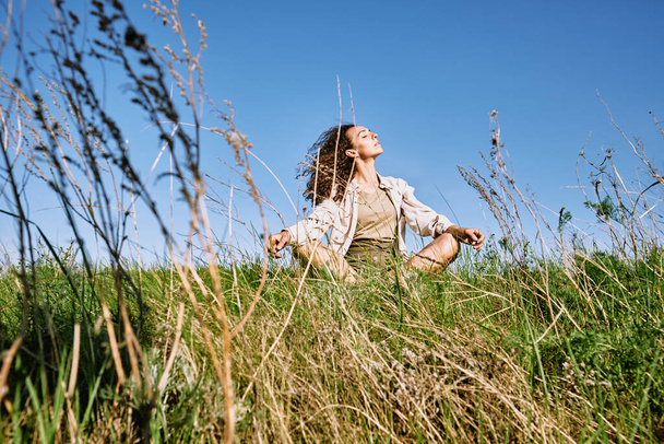 Νεαρή χαλαρή γυναίκα με σκούρα μακριά κυματιστά μαλλιά σταυρώνουν τα πόδια, ενώ κάθεται στο πράσινο γρασίδι κατά του μπλε ουρανού και απολαμβάνοντας ανάπαυση την ηλιόλουστη μέρα - Φωτογραφία, εικόνα