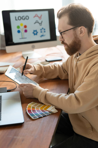 Koncentrált szakállas férfi kapucnis pulóverben ül a fa asztalnál, és táblagépet használ, miközben a márka tervezésén dolgozik - Fotó, kép