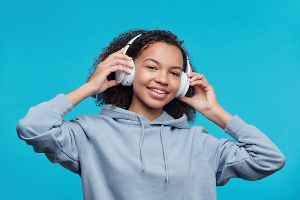Retrato de menina preta bonita feliz com capuz ajustando fones de ouvido sem fio enquanto ouve música contra fundo azul - Foto, Imagem