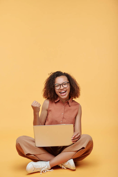 Portrait of excited African-American student girl sitting with crossed legs on floor and making yes gesture while studying online course on laptop - Photo, Image