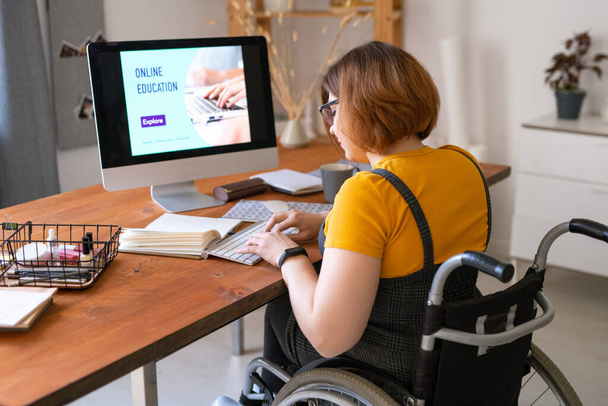 Jonge vrouw in rolstoel drukken toetsen op het toetsenbord in de voorkant van de computer monitor, terwijl gaan om remote les in huis omgeving - Foto, afbeelding