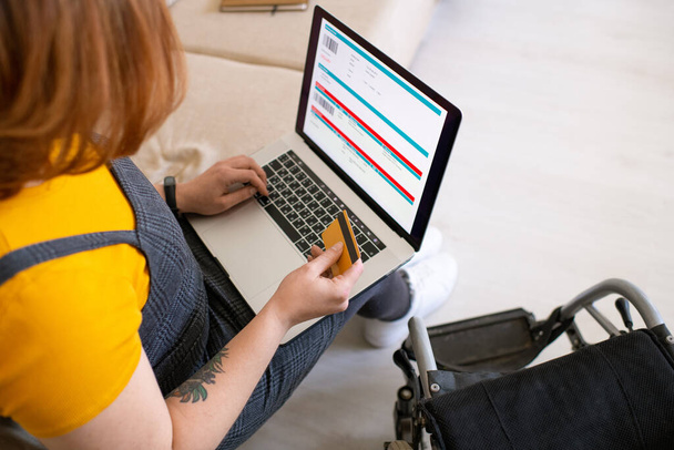 Молодая женщина-инвалид с кредитной картой над клавиатурой ноутбука, сидящая на диване, пока собирается забронировать и оплатить авиабилеты из дома - Фото, изображение
