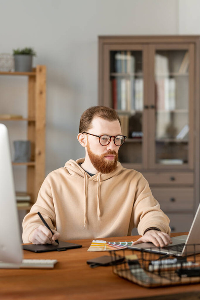 Gözlüklü, ciddi bir sakallı adam grafik tabletine tasarım çizimi çiziyor ve dizüstü bilgisayarda düzenliyor. - Fotoğraf, Görsel