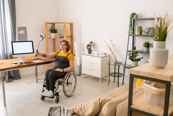 Mladá zdravotně postižená žena blogger v invalidním vozíku držet hůl s chytrým telefonem ve fontu na sebe při nahrávání videa pro její on-line publikum - Fotografie, Obrázek