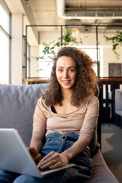 Portret uśmiechniętego młodego menedżera SMM z kręconymi włosami siedzącego na wygodnej sofie i pracującego z laptopem w kawiarni - Zdjęcie, obraz