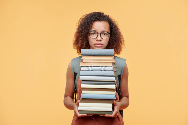 Upset frowning Afro-American girl with curly hair holding pile of books against yellow background, time to prepare for exam concept - Photo, Image