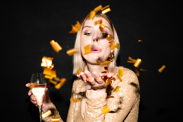 Νεαρή όμορφη γυναίκα με ξανθά μαλλιά με φλάουτο σαμπάνιας φυσάει χρυσά κομφετί από το χέρι της ενώ απολαμβάνει το πάρτι - Φωτογραφία, εικόνα