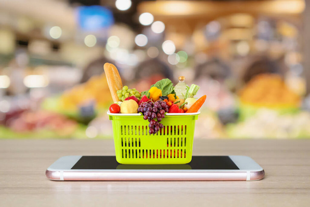 Fresh food and vegetables in shopping basket on mobile smartphone on wood table with supermarket aisle blurred background grocery online concept - Photo, Image