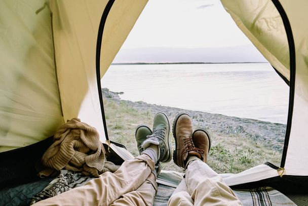 Poten van jong amoureus echtpaar in casualwear liggend in open tent in de voorkant van de kust en ontspannen tijdens het wandelen tijdens het zomerweekend - Foto, afbeelding