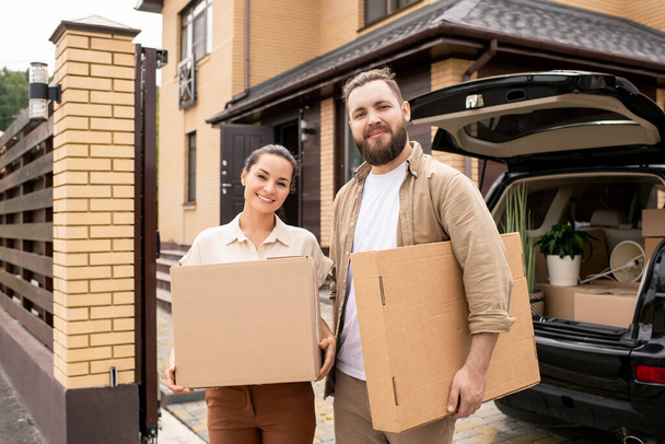 Portrait of smiling young couple standing at open car trunk and holding moving boxes against new house - Photo, Image