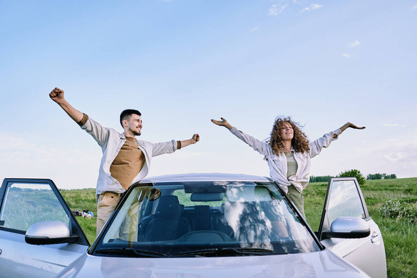 Ευτυχισμένος νεαρός άνδρας και γυναίκα με απλωμένα τα χέρια να στέκονται δίπλα στις ανοιχτές πόρτες του αυτοκινήτου τους και να απολαμβάνουν ζεστή καλοκαιρινή μέρα ενάντια στον γαλάζιο ουρανό - Φωτογραφία, εικόνα