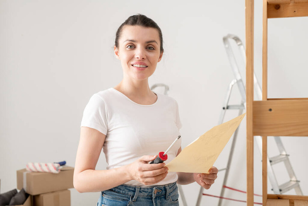 Porträt einer lächelnden attraktiven jungen Frau, die einen Schraubenzieher in der Hand hält und Möbel nach Anleitung zusammenbaut - Foto, Bild