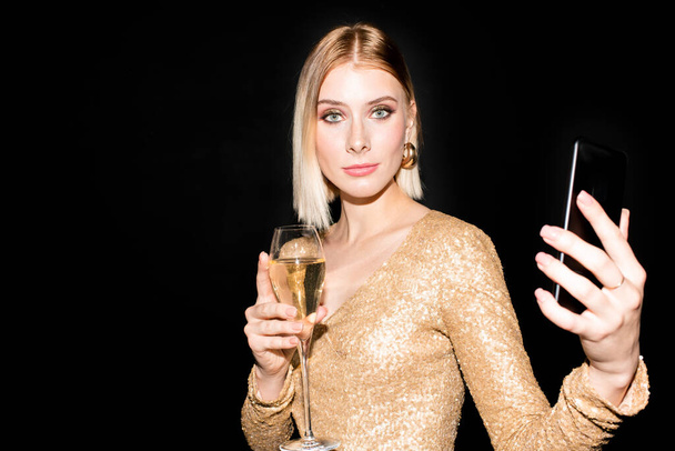 Young pretty blond woman looking at you while holding smartphone and flute of champagne and and enjoying birthday or new year party - 写真・画像