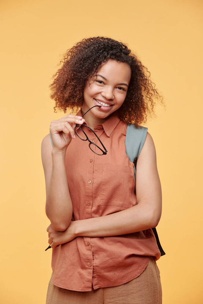 Portrait of positive black girl with curly hair holding eyeglasses against bright background - Photo, Image